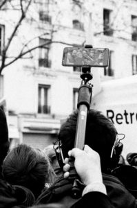 A black-and-white photo of a crowd of people, one holding a selfie stick with a smartphone at the top. The phone screen displays a larger crowd scene from the manifestation on 11 Janvier 2020 against the réforme des retraites. The background features an out-of-focus building with windows and faint tree branches.