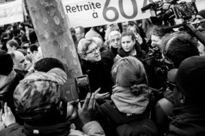 A black and white photo captures a manifestation scene on 11 Janvier 2020, with a man addressing a crowd surrounded by cameras and microphones. A banner behind them boldly displays "Retraite à 60" as people hold up their phones to capture the moment.