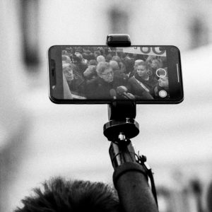 A black and white image shows several people speaking into microphones, captured on a smartphone mounted on a tripod or hand-held stick. The scene, recorded during the manifestation against the réforme des retraites on 11 Janvier 2020, displays a close-up of a person speaking in front of others.