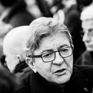 Black and white close-up photo of a middle-aged man with gray hair and glasses, taken during the réforme des retraites manifestation on 11 janvier 2020. He looks slightly away from the camera, surrounded by blurred figures in the high-contrast background, capturing his detailed expression.