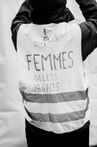 A black-and-white photo of a person adjusting their hood while wearing a reflective vest with handwritten text that reads "FEMMES GILETS JAUNES". The vest features an illustration of the Eiffel Tower and two small triangle shapes, marking the 11 Janvier 2020 manifestation against the réforme des retraites.