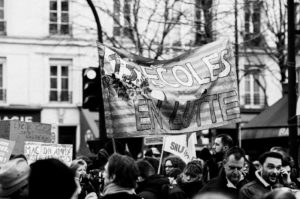 A black and white photo of a crowded street protest. People hold signs and banners, including a prominent one reading "15 écoles en lutte" (15 schools in struggle). The background shows buildings and leafless trees, indicating it might be autumn or winter, possibly during the réforme des retraites manifestation on 11 Janvier 2020.