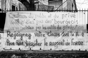 A large banner is displayed on a fence with handwritten French text urging the public and private sectors to unite and take action against the réforme des retraites. The banner, dated 11 Janvier 2020, is partially covered with transparent plastic, set against an outdoor area with a brick building and leafless tree in the background.