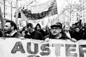 A crowd of protestors marches down a street, holding various signs and banners. The central banner reads "ÉCOLES EN LUTTE" (Schools in struggle), referencing the réforme des retraites. Another prominently displayed banner has the word "AUSTER." The protestors, gathered on 11 Janvier 2020, appear focused and determined.