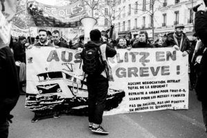 A black and white photo shows a group of people marching with a large banner reading "AUSTERLITZ EN GRÈVE !" and additional protest messages. One person in the foreground holds a megaphone while others carry signs and banners, part of the 11 Janvier 2020 manifestation against the réforme des retraites.