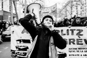 A black and white photo of a protest on 11 Janvier 2020. A person is in the foreground, wearing a beanie and holding a megaphone while speaking into it about the réforme des retraites. Behind them, a crowd holds a large banner with text and images, as well as hand-drawn illustrations of cars. Trees and buildings are visible in the background.