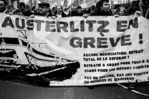 A black-and-white photo shows a group of protesters holding a large banner that reads "Austerlitz en Grève!" Their demands include "Aucune négociation, retrait total de la réforme des retraites." People march in the background during the manifestation on 11 janvier 2020.