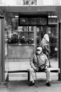 A black and white photo shows an older man sitting at a bus stop, wearing a hat, sunglasses, and holding a cane. Behind the glass of the shelter is the reflection of another person, possibly from a manifestation on 11 Janvier 2020. The scene is in an urban setting, with a building visible in the background.