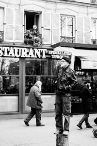 Black-and-white photo of a street scene. A man, perhaps part of the réforme des retraites movement of 11 Janvier 2020, stands balanced on a post, wearing a leather jacket and beret. Behind him, pedestrians walk by a storefront with the word "RESTAURANT" visible. A person is looking out from a window above the restaurant.