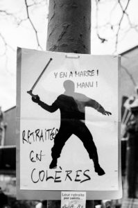 A poster is attached to a pole, depicting a silhouette of a person holding a baton. The text reads "Y EN A MARRE! MANU!" and "RETRAITÉS EN COLÈRES," translating to "Fed up! Manu!" and "Retirees Angry." This manifestation against the réforme des retraites took place on 11 Janvier 2020, with a blurred city
