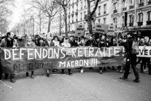 A black-and-white photo of protesters marching on a street, holding a large banner that reads "DÉFENDONS NOS RETRAITES! MACRON!!!" during the manifestation on 11 Janvier 2020. Other signs related to the réforme des retraites are visible. The background shows tall buildings and leafless trees, with a serious and determined mood.
