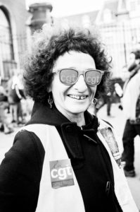 Black and white photo of a smiling person with curly hair wearing heart-shaped patterned sunglasses and a vest with a "CGT Centrale Finances" sticker. The background features blurred buildings and people, indicating a street setting during the 11 Janvier 2020 manifestation against the réforme des retraites.
