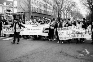 A black and white photo shows a large group of people marching on a street, holding banners and signs. The banners indicate they are protesting about the RER B train line and calling for retirement benefits. Leafless trees and buildings loom in the background, marking the manifestation of 11 Janvier 2020.
