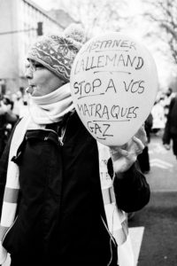 A person in a winter hat and scarf holds a balloon with a handwritten message in French that reads, "Castaner Lallemand stop à vos matraques gaz". The person is blowing a whistle, surrounded by a crowd during the 11 Janvier 2020 manifestation against the réforme des retraites.