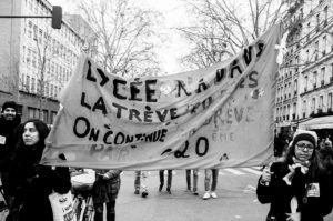 Black and white photo of a group of people holding a large, hand-painted banner during a street manifestation. The banner reads "Lycée Raspail, La grève aux élèves, On continue la grève, Paris 20ème." They are walking under leafless trees in an urban area, protesting the réforme des retraites.
