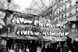 A black-and-white photo of a 11 Janvier 2020 street protest where people are holding a large banner with graffiti-style text. Trees and a multi-story building line the background, highlighting the manifestation against the réforme des retraites. A "METRO 8" sign is visible on the right side.