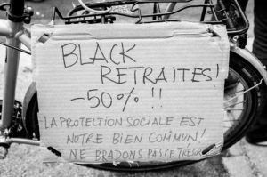 A cardboard sign attached to the back of a bicycle with a message written in French: "BLACK RETRAITES! -50%!! La protection sociale est notre bien commun! Ne bradons pas ce trésor!" The sign, reminiscent of the manifestation on 11 Janvier 2020, advocates for the protection of social security.