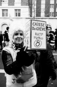 A person wearing a curly wig and gloves holds up a sign that reads "CAISSE DE GRÈVE RATP METRO 8" at a manifestation on 11 Janvier 2020. The background shows other protesters and buildings. The image is in black and white.