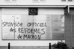 A black and white image captures graffiti on a shopfront's blank metal panels, reading "SPONSOR OFFICIEL des REFORMES de MACRON" in French. The shop sign above reads "LCL," indicating a bank. This was taken during the manifestation on 11 Janvier 2020, addressing the réforme des retraites.