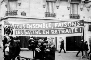 A black-and-white photo of a street protest in front of a building on 11 Janvier 2020. Demonstrators hold a banner in French that reads, "TOUS ENSEMBLE, FAISONS-LES BATTRE EN RETRAITE!" People, including a cameraman and a woman reading a document about réforme des retraites, are gathered around.