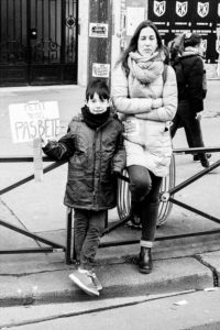 A black-and-white photo shows a woman and a child standing on a sidewalk during the manifestation of 11 Janvier 2020. The child holds a sign reading "Petit mais pas bête." The woman is leaning against a fence with her arms crossed. Both are dressed warmly in coats and scarves. People and posters are visible in the background.
