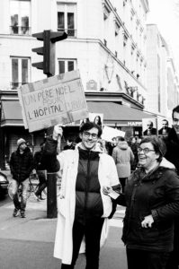 A person in a white coat holds a sign reading "Un Père Noël pour l'hôpital public" ("A Santa Claus for the public hospital") at what appears to be a manifestation on 11 Janvier 2020. Another person stands beside them smiling. The setting is a city street, likely amidst protests over the réforme des retraites.