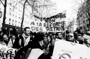 Black and white image of a lively street protest on 11 Janvier 2020. Demonstrators hold banners, one of which says "RATP en greve non à la réforme des retraites," opposing pension reforms. The crowd is diverse, with people wearing jackets and scarves, and a few trees are visible behind.