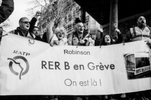 A group of people wearing jackets and scarves enthusiastically hold up a sign in French that reads "Robinson RER B en Grève, On est là !", indicating a transit strike on the RER B line. They appear to be protesting against the réforme des retraites, with some raising their fists in the air during this manifestation on 11 Janvier 2020.