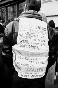 Black and white photo of a person wearing a jacket decorated with paper that has French writing on it. The writings express strong opposition to "egoists" and "exploiters" and emphasize a demand for liberty, equality, and solidarity amongst people, reflecting the spirit of the manifestation on 11 Janvier 2020.