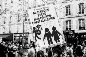 A black-and-white photo shows a crowded street protest. In the foreground, a protester holds a large sign that reads, "A cause de Macron, BlackRock prend mon pognon: capitalisation." The sign depicts a figure taking money from another figure holding a cane. This manifestation took place on 11 Janvier 2020 amid the réforme des retraites debate.