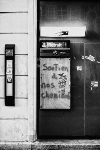 Black and white photo of a boarded-up entrance with graffiti. The graffiti reads, "soutien à nos chemin" with a doodle of a face. Nearby, a panel has posters including one with "ART EN GREVE," pinned to the wall during the manifestation against réforme des retraites on 11 Janvier 2020.