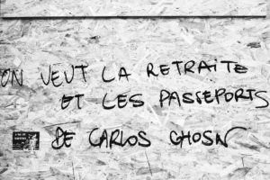 A grayscale image of a wall with graffiti in French reading "ON VEUT LA RETRAITE ET LES PASSEPORTS DE CARLOS GHOSN," referencing the réforme des retraites. A small sticker on the left depicts a figure with a microphone and text "A BAS LES FRONTIÈRES !" (Down with borders!). The wall has a rough, textured surface.