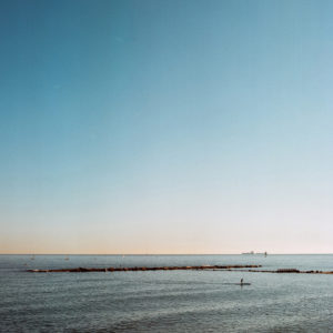A serene ocean scene off Barceloneta with a lone paddleboarder gliding over calm waters. A distant line of rocks stretches horizontally, and the horizon is visible under a clear blue sky. Sailboats and a large ship are seen far away near the horizon, completing this tranquil view of Barcelona's coastline.