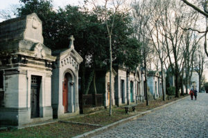A cobblestone path lined with old mausoleums in Père Lachaise Cemetery. Trees without leaves surround the area, and two people walk in the distance. The mausoleums have various architectural styles and are covered with light vegetation. The atmosphere is serene and solemn.