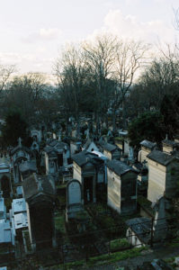 An aerial view of the Père Lachaise Cemetery reveals numerous tombstones and mausoleums. The trees are bare, indicating a winter or early spring season. The sky is partly cloudy, and the scene is serene and somewhat melancholic.