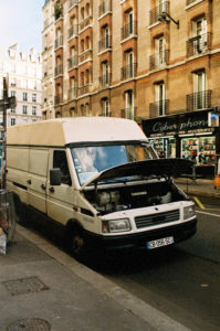 A white delivery van, appearing to be in panne with its hood open, is parked on a city street. Behind the camionnette, a shop named "Cyber phone" and a multi-story building with balconies are visible. The scene is captured during daylight, and pedestrians can be seen in the background.