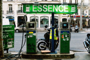 A person in a yellow vest refuels a motorcycle at an urban gas station, illuminated by a neon-lit "Essence" sign. The station offers two pumps for diesel and SP 95. In the background, buildings, parked cars, and scooters line the bustling street.