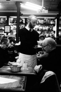 A black and white image of a waiter serving an elderly man at Le café d'Hector. The waiter is holding a tray, and the elderly man is reaching for a napkin. Several other patrons are seated in the background, and shelves filled with bottles and products are visible.