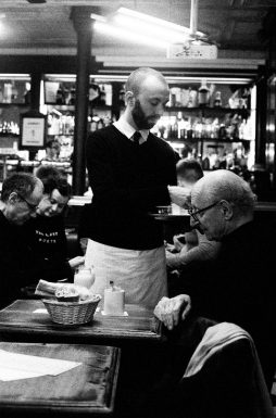 2020-02-01-Canon-EOS-30V-Kodak-TMAX3200_11_MOD_BW_FRAME_resize A black and white image of a waiter serving an elderly man at Le café d'Hector. The waiter is holding a tray, and the elderly man is reaching for a napkin. Several other patrons are seated in the background, and shelves filled with bottles and products are visible.