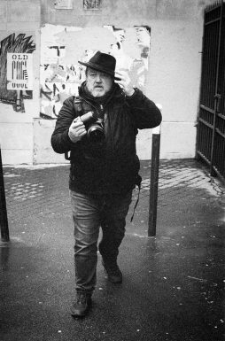 2020-02-01-Canon-EOS-30V-Kodak-TMAX3200_35_MOD_BW_FRAME_resize A person wearing a hat and coat walks down a wet street while holding a camera. The background features a wall with posters, one of which says "OLD LOVE OUVL." The street is bordered by a metal fence on the right and a pole near the person. Captured by Marc Lepage, the image is in black and white.