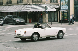 A white vintage convertible car is being driven on a city street. The background shows a cafe with striped awnings and a restaurant with a large movie poster. The two passengers in the classic car are bundled up, suggesting chilly weather.
