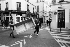Two people are engaged in the transport de canapé, carrying a large sofa down a narrow street in an urban setting. Surrounded by buildings with shops like "elevenparis," pedestrians and a cyclist can be seen in the background. The scene appears to be candid, capturing everyday life with attention to detail.