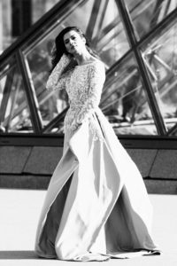 A black-and-white photo captures a woman wearing an elegant, long-sleeved lace gown with a flowing skirt, beautifully epitomizing la belle robe. She stands in front of an abstract, geometric architectural structure, slightly tilting her head and touching her hair.