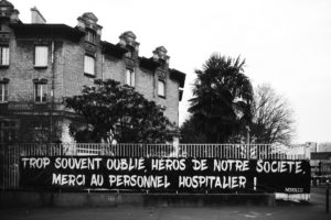 A black and white photograph of a building with a large banner on the fence in front. The banner reads in French, "Trop souvent oubliés, héros de notre société, merci au personnel hospitalier!" Trees and bushes are visible behind the fence.