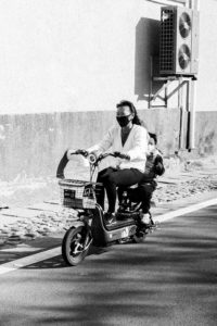 A black-and-white photo captures a person riding a motorized scooter with a basket on the front. They are wearing face masks, and a child sits behind them, also masked. The backdrop reveals a textured wall and air conditioning units mounted high, reminiscent of scenes along the route de Mutianyu in China.
