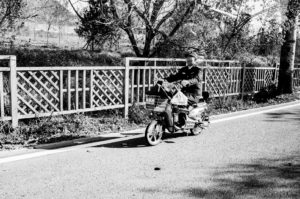 A black-and-white photo shows a person riding a bicycle-like moped on the route de Mutianyu in Chine on a sunny day. The rider, wearing a cap and coat, has a basket with items attached to the front of the moped. They are traveling along a paved road with a lattice-fenced area in the background.