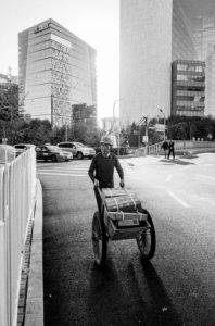A person pushes a charrette à bras loaded with packages along a sunlit city street in Pékin, Chine. Tall, modern buildings with glass facades tower in the background. Cars are parked by the roadside, and a few pedestrians are visible in the distance. The image is in black and white.