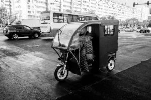 A black and white photo of a small three-wheeled vehicle, likely a rickshaw, at a busy intersection in Pékin. A person is seated inside the motos-like vehicle, which has a covered cabin. Several cars and a bus are in the background, with buildings lining the street.