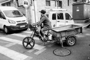 Black and white image of a person riding a motorized tricycle with a cargo bed, crossing a street at a pedestrian crosswalk in Pékin. The scene includes parked motos, a pedestrian crossing sign, and brick buildings in the background.