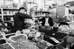 A black and white image of a bustling indoor marché in Pékin showcases a stall selling a variety of nuts and dried fruits. Two men are engaged in a transaction: one man is filling a plastic bag with nuts while the other looks on. Various containers and jars of products are displayed, typical of markets in Chine.